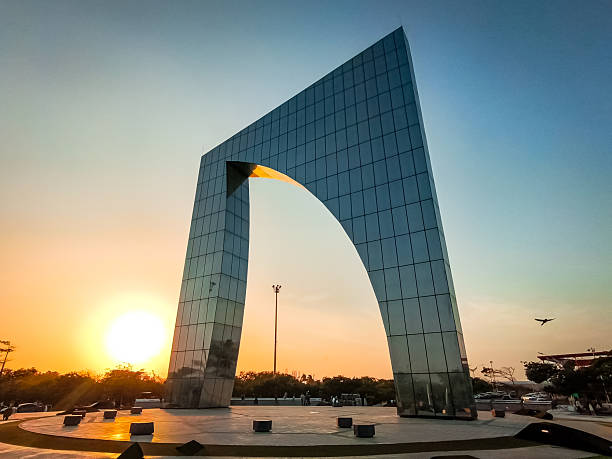 Ventana de campeones, Barranquilla, Sleek modern glass arch monument standing in a city plaza at sunset, with warm golden light, reflective surfaces, and a clear evening sky.